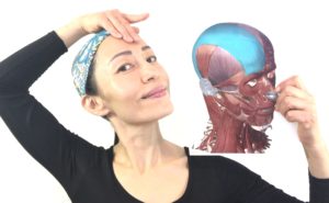 A woman with a blue cap touching her forehead and smiling, while holding a photo showing the musculature of human face.