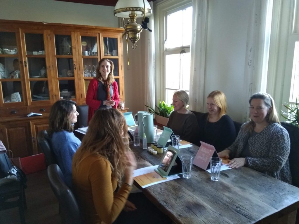 After Face Yoga Teacher Certification: Viktorija standing at a table and smiling while teaching a class of 5 other women sitting at the table. 