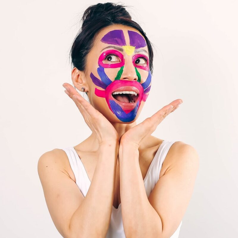 Fumiko Takatsu smiling while looking up with a colorful - blue, purple, green, pink and yellow - facial mask