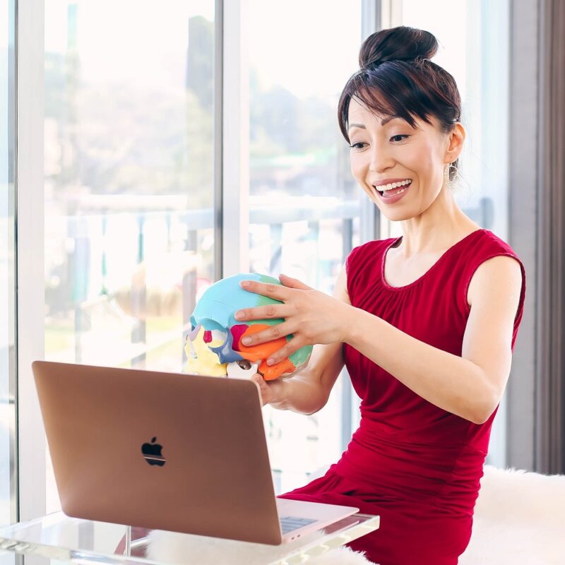 Fumiko Takatsu smiling and holding a colorfully painted skull and pointing it to the laptop screen while teaching Face Yoga online.