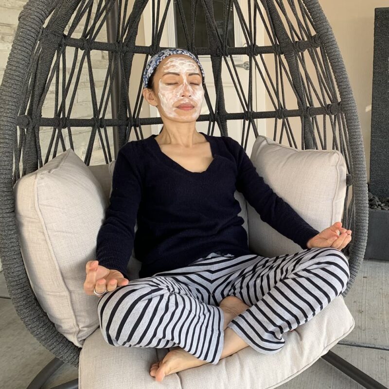 Fumiko Takatsu sitting with closed eyes, legs crossed in a gray egg chair with a white facial mask on her face.
