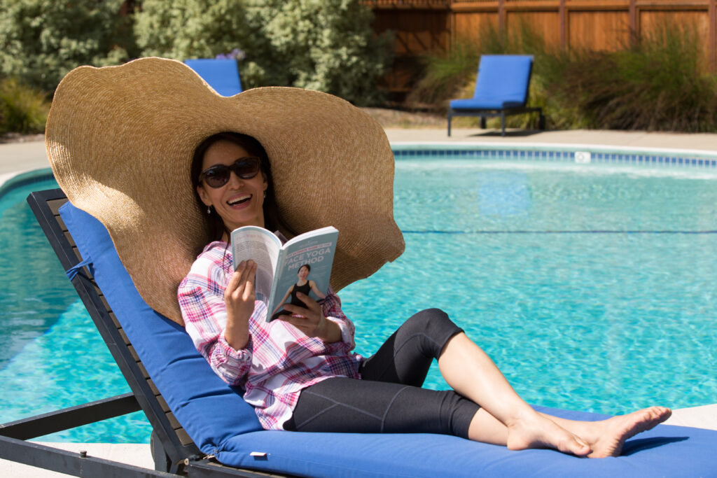 Fumiko Takatsu, a renowned Face Yoga instructor, sitting at the pool with a hat and reading a book.  