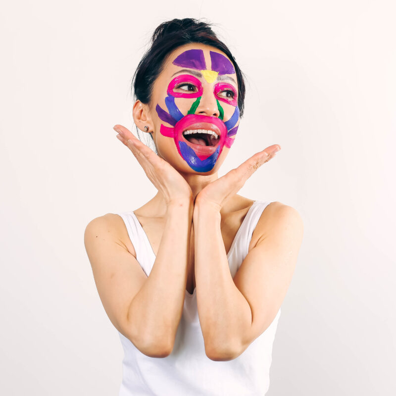 Fumiko Takatsu smiling while looking up with a colorful - blue, purple, green, pink, and yellow - facial mask.