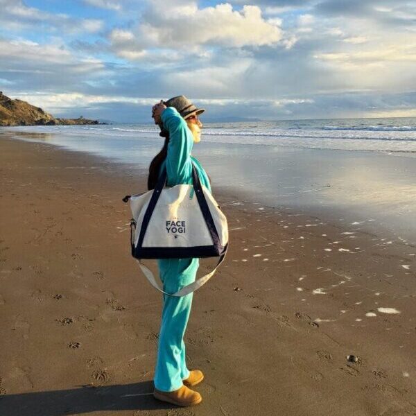 A woman in green clothes with a duffel bag on her shoulder, standing on the sand beach and looking at the ocean.