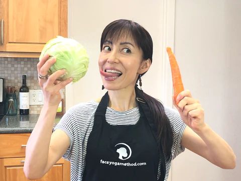 A woman wearing apron, holding cabbage and carrot, in a kitchen